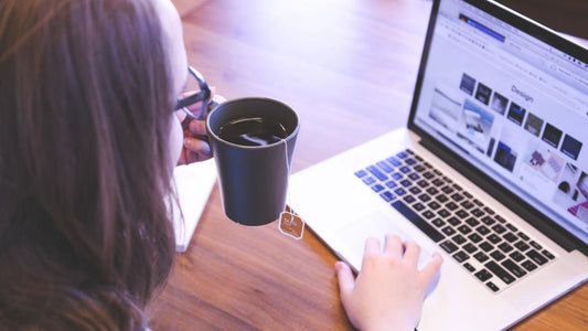Person at a desk drinking tea while browsing on a laptop, seen from behind