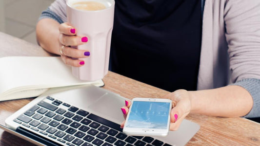 Hands holding a phone and a mug over a laptop and notebook